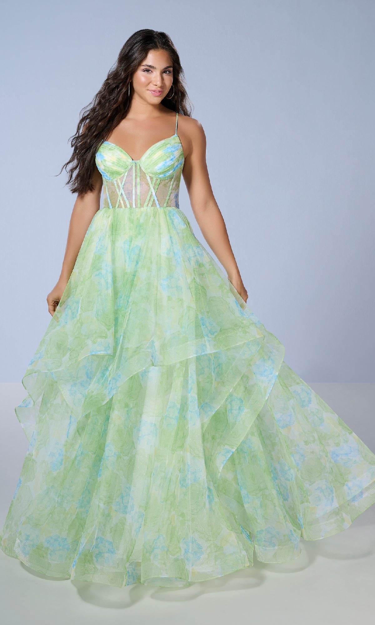 A woman stands against a plain background wearing the Long Prom Dress: Tiffany 16163, featuring a light green and blue floral-print design with a fitted sheer corset bodice and spaghetti straps.